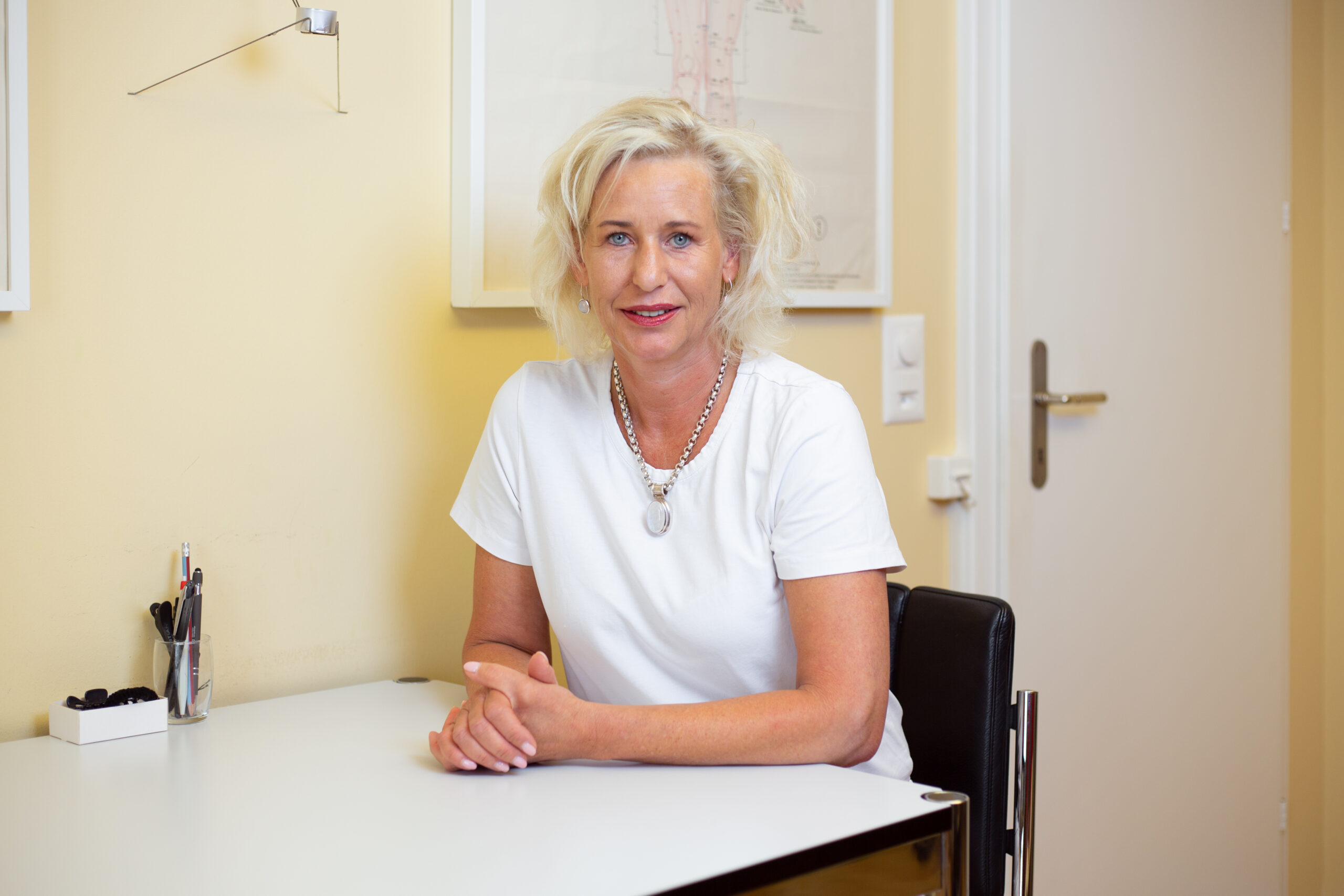 TCM-Gynäkologin Brigitte Weber sitzt an einem Tisch vor einer gelben Wand in ihrer Praxis.