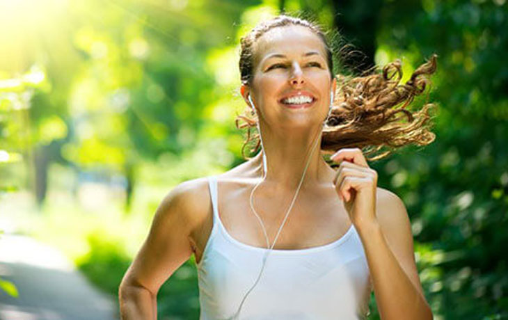 Eine junge Frau mit Kopfhörern im Ohr joggt an einem sonnigen Tag in einem weissen, ärmellosen T-Shirt an grünen Bäumen vorbei.