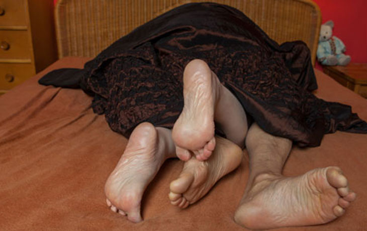Das sind die Füsse von einem Mann und einer Frau, die unter einer leichten, dunkelbraunen Decke in einem Bett liegen